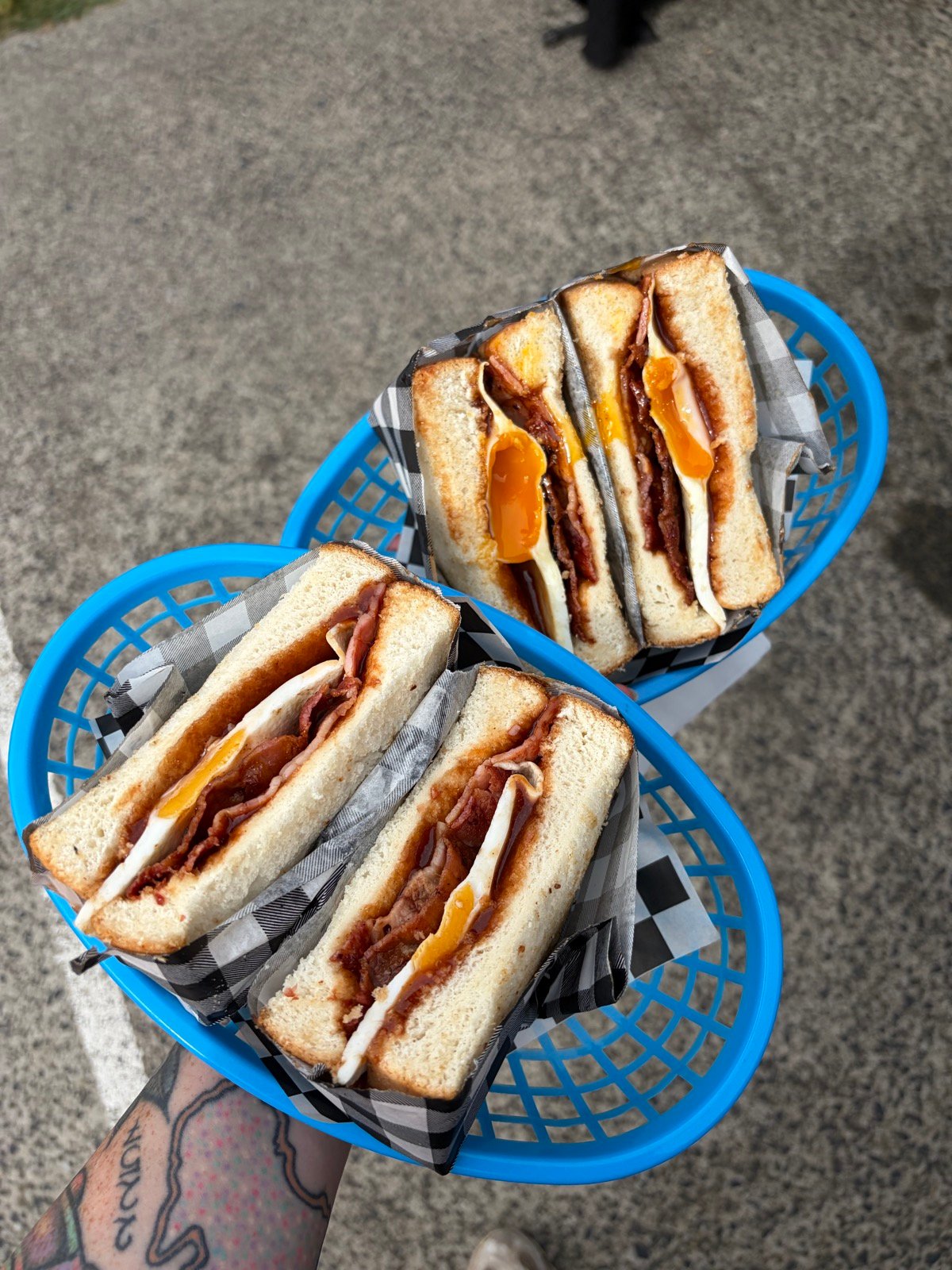 Two bacon-and-egg toasties on gingham paper in blue baskets, held over a concrete kerb by a tattooed arm.