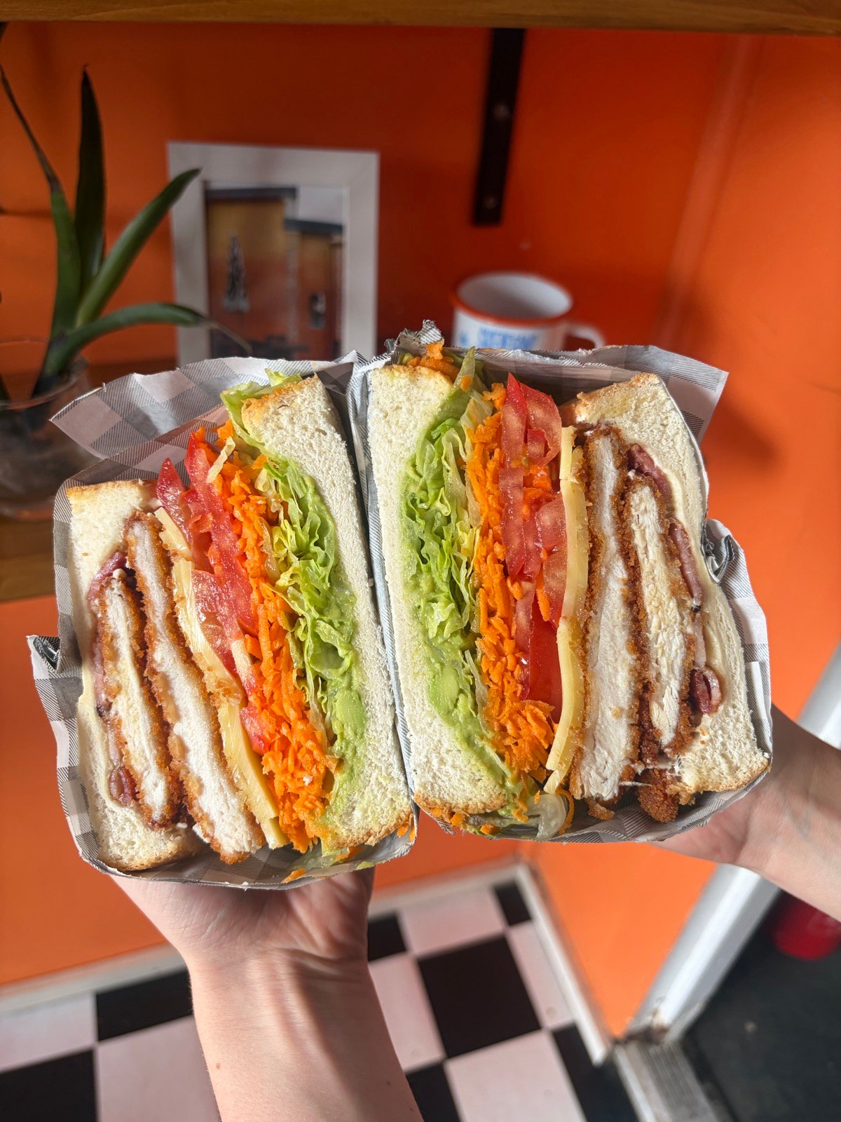 Two schnitzel sanga halves held up cross-section — crumbed chicken, cheese, lettuce, tomato and grated carrot on thick white bread, orange wall behind.